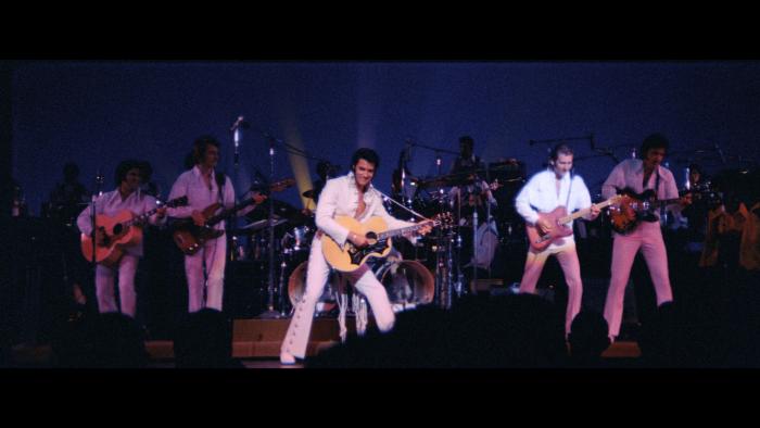 EPiC: Elvis Presley in Concert movie: Presley performs with a wink and a smile, holding his guitar to the right with his band members similarly posed