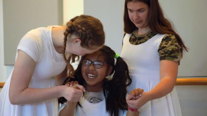 Three girls in white dresses holding hands while the one in the middle smiles broadly.