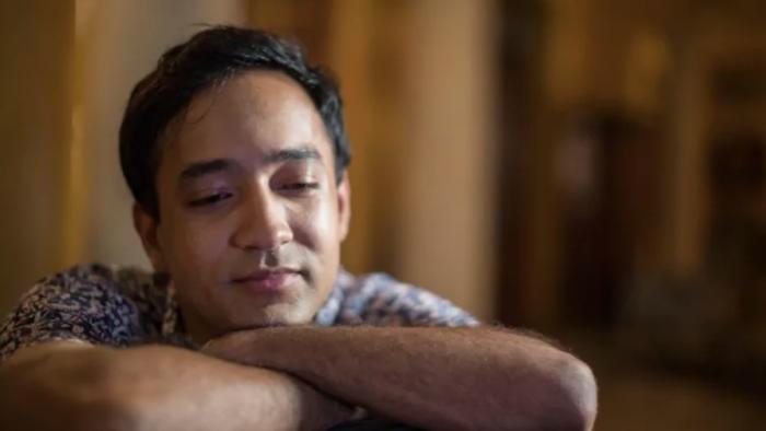 Funny Boy Movie: A man smiles fondly, gaze downward as he rests his chin on his arms