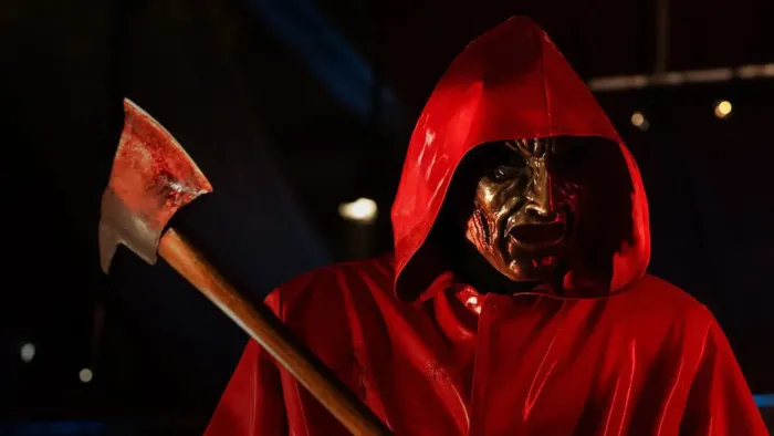 Fear Street: Prom Queen movie: A killer in red hoodie and face mask right wields a bloody axe left while looking for victims outside at night