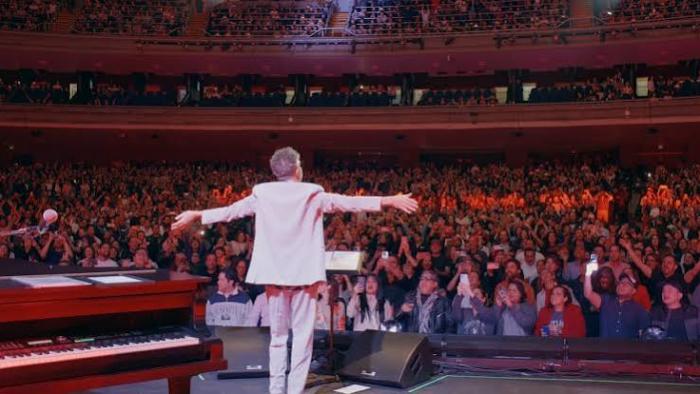 Fito Páez in white facing cheering audience in a large theater.