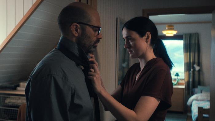Fjord: A woman adjusts her husband's tie as they both look serious standing in the hallway of their home