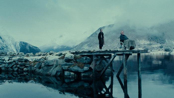 Fjord: A woman stands behind a man, who sits in a wheelchair at the end of a peer, overlooking a lake surrounded by snowy mountains