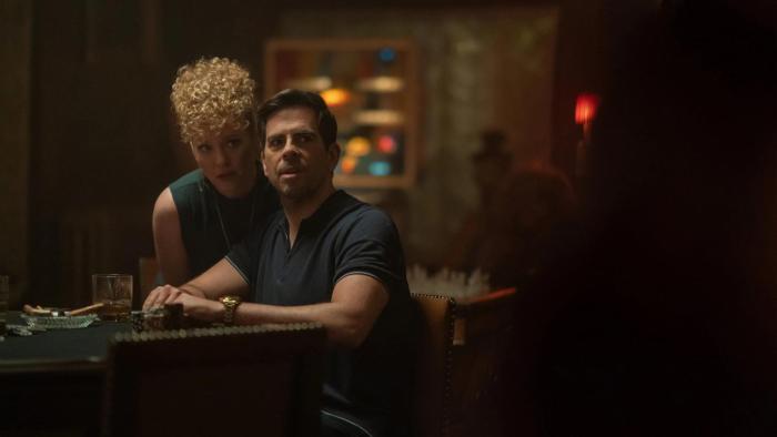 Night Always Comes: A man looks at someone off camera as he sits at a table, a woman standing behind him