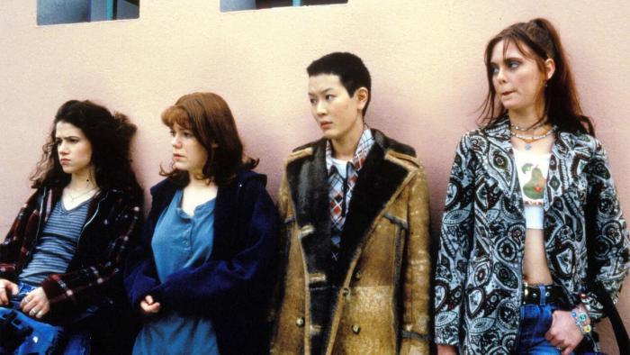 Foxfire: Four teenage girls in varied outfits stand against a pink wall, appearing thoughtful