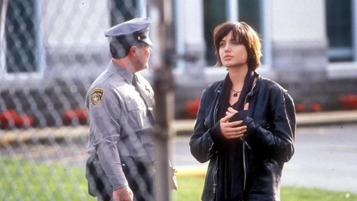 Foxfire: Woman in a leather jacket walks out of a fenced institution as a security guard looks on
