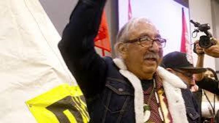 Free Leonard Peltier Movie: Older man with glasses and raised arm at event, flags in background.