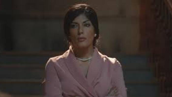 From the Ashes Movie: Woman in pink suit and pearls looks serious