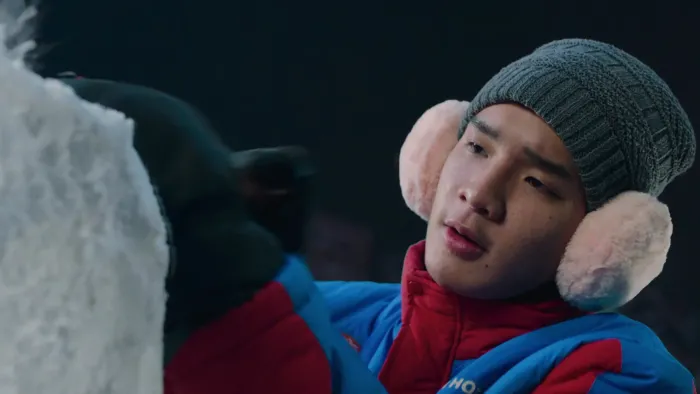 Frozen Hot Boys movie: Thai older teen in red and blue coat, hat, and pink earmuffs, right, works on ice sculpture outside at night 