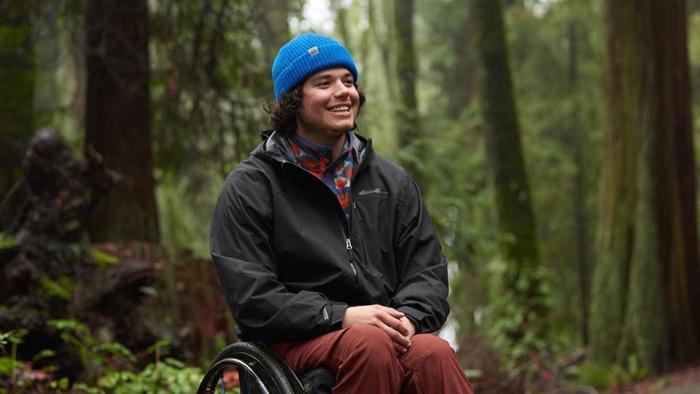 Still from Full Circle: White professional sit skier Trevor Kennison sits in a wheelchair in the woods, wearing a blue ski cap, a red, white, and blue fleece sports shirt, black Eddie Bauer windbreaker jacket, brown pants, and black sport shoes