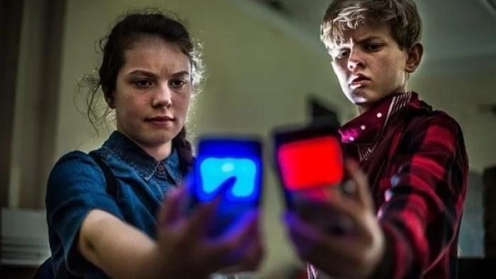 Future TX Movie: Molly and Dylan hold their futuristic cell phones at arm's length; the devices are glowing blue and red