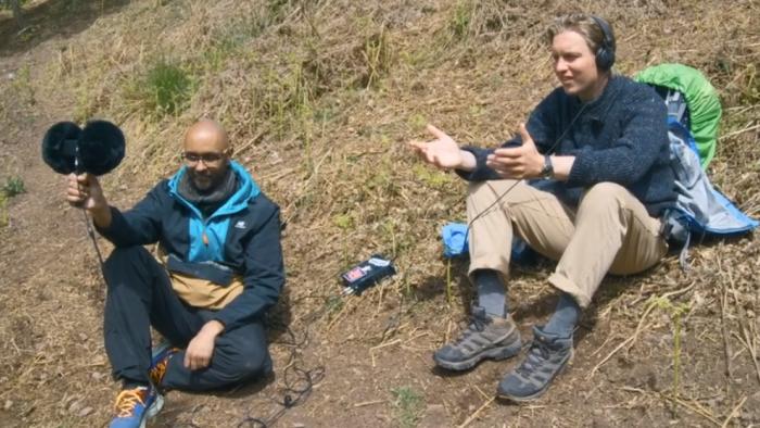 George Ezra -- End to End: George Ezra and another man sit on hillside listening to binaural sounds with microphone and earphones