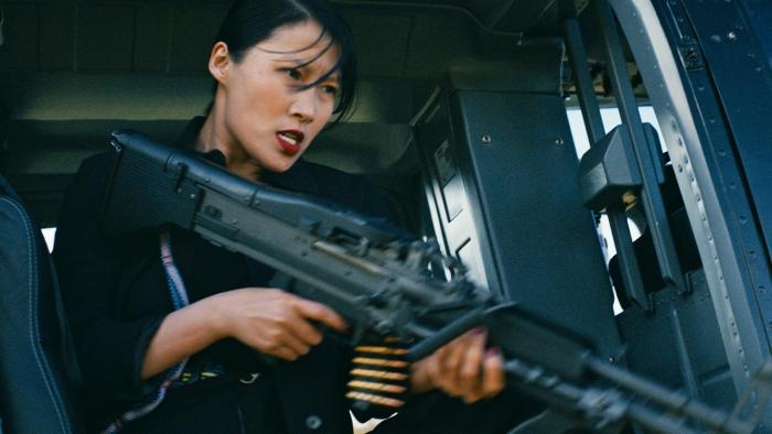 Get Fast movie: Chinese Canadian woman in black center left looks out the side of a helicopter while holding a large machine gun 