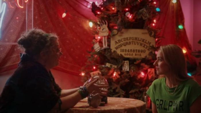 Ghosts of Christmas Past: A young White blond woman sits in a festive tent in front of a fortune teller with a snow globe for a crystal ball.