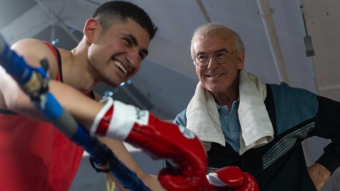 Giant: Young boxer smiling with a coach, leaning on boxing ring ropes