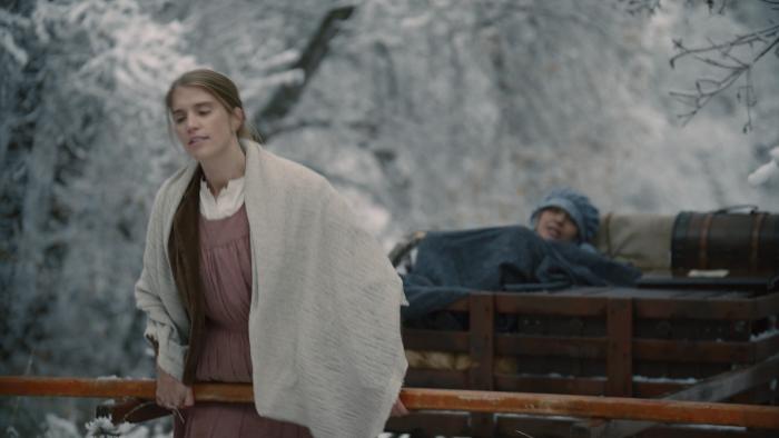 Go West Movie: White woman in prairie garb walks through a snowy trail carrying a handcart with another woman lying in it