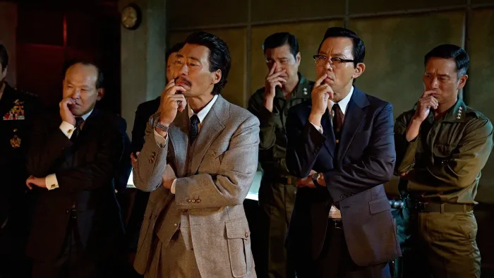 Good News movie: Five Korean men in dark colored suits all have one of their hands to their chins, the other hand across waist, all looking puzzled