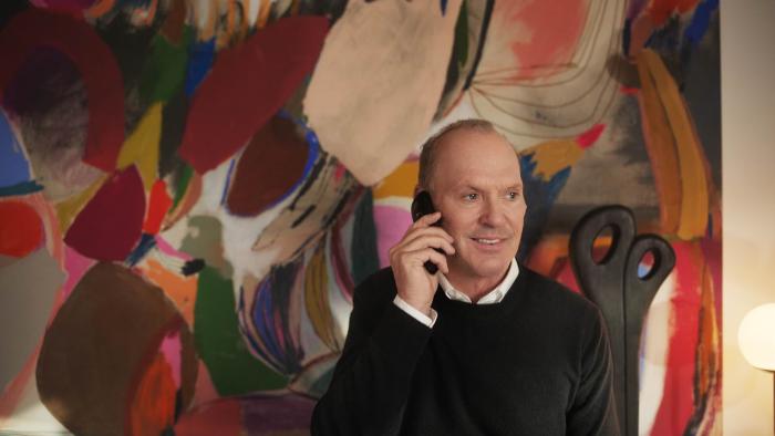 Goodrich Movie: Smiling Andy Goodrich (Michael Keaton) speaks on the phone, in front of a colorful work of art