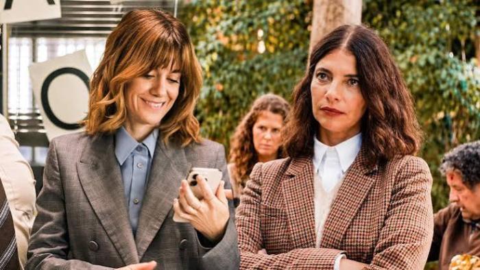 Go Team! Movie: Two women in business attire, one smiling at a phone, the other with crossed arms.