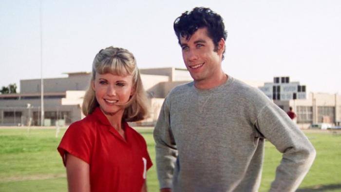 Grease Movie: Olivia Newton-John in a red polo shirt stands next to John Travolta in a gray sweatshirt outside on a high school field