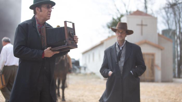 Gunslingers Movie: Ben (Nicolas Cage), wearing a hat and dark glasses, holds an old-fashioned camera; another man watches him