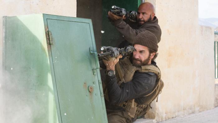 Guy Ritchie's The Covenant Movie: Ahmed (Dar Salim) and Sergeant John Kinley (Jake Gyllenhaal), in civilian clothes, take cover behind a small metal d