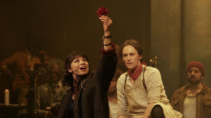 Hadestown the Musical: Eurydice (Eva Noblezada) holds a red flower up and sings while Orpheus (Reeve Carney) looks on