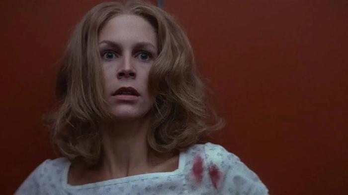 Halloween II: Jamie Lee Curtis looks scared as she stands against a red wall wearing a hospital gown with blood on the shoulder