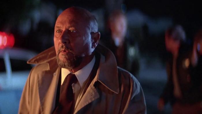 Halloween II: Donald Pleasance looks serious as he stands in a street with policemen and police cars in the background