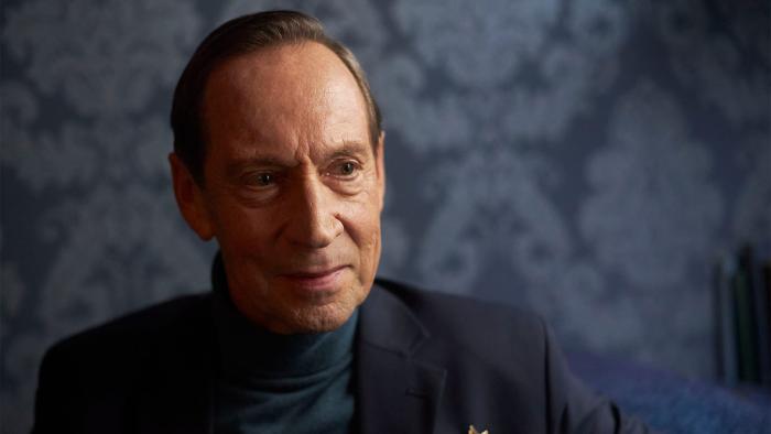 Hamlet: Jonathan Hyde smiles slightly against a background of blue patterned wallpaper