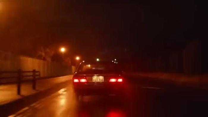 Hard Days movie: Back of a black car center drives down the street at night in the rain with a streetlamp on left