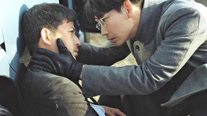 Hard Days movie: Japanese man in grey coat, glasses, and black gloves on right holds Japanese man's neck while he is slumped on ground on left leaning against a white car