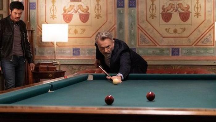 Assassin Club: An older White man is about to play a shot in pool while a younger Asian man looks on