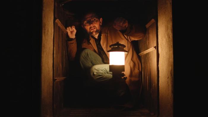 Hokum Movie: Ohm Bauman (Adam Scott) crouches in a dumbwaiter, holding a lantern up to the darkness
