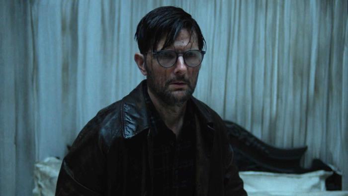 Hokum Movie: Wet and covered in dark gunk, Ohm Bauman (Adam Scott) is in a grimy hotel room looking scared