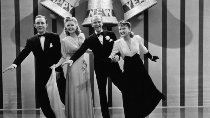 Holiday Inn Movie: Four White adults in suits, bowties, and formal gowns stand on stage in front of large bells that read "Happy New Year"