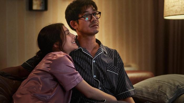 Home for Rent movie: Thai woman in pajamas left sits on couch hugging Thai man in pajamas center in darkly lit living room