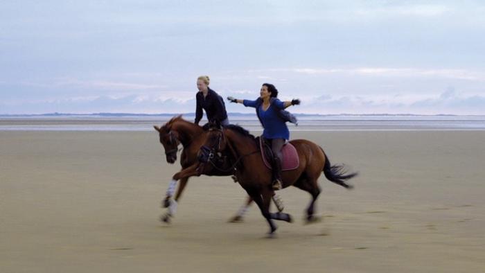 Of Girls and Horses Movie: Two people riding horses on a beach, arms outstretched, cloudy sky.