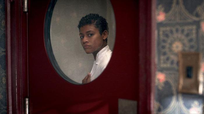 House of Spoils movie: Black woman with short black hair and in white coat looks through circle window on red door to kitchen