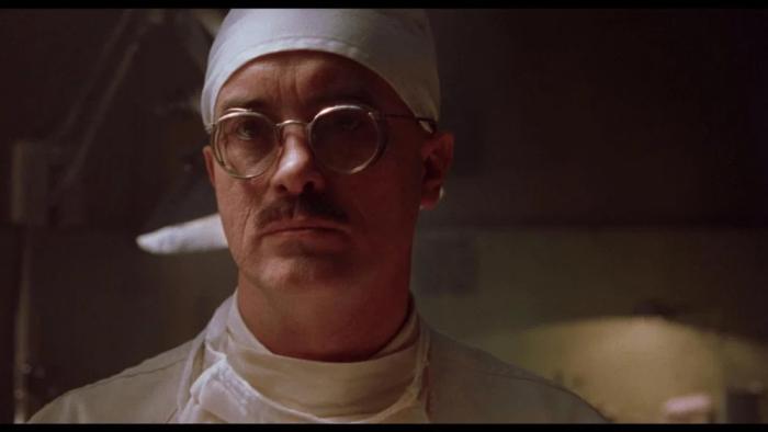 House on Haunted Hill: Dr. Vannacutt in old-fashioned surgical scrubs in a dark room looking serious