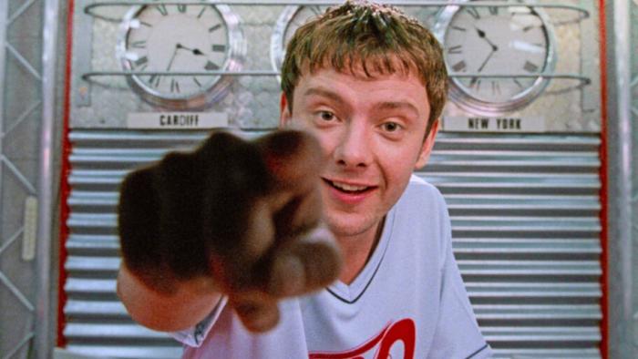 Human Traffic: A young White man points down the camera at the viewer with three wall clocks behind him