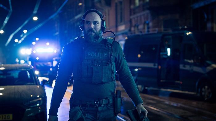 iHostage movie: White Dutch bald man in full police gear, headset, and bullet proof vest holds tablet walking down city street at night