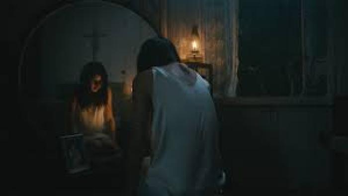 In My Mother's Skin movie: Filipino woman center in white negligee hunches in front of mirror in dark bedroom with reflection showing a cross above her head
