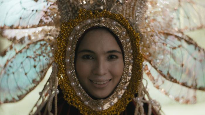 In My Mother's Skin movie: Filipino woman center dressed elaborately in bejeweled mask and headdress that looks like a flower surrounding her face smiles in front of window