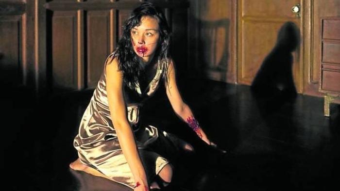 In My Mother's Skin movie: Filipino woman in white negligee center sits on her legs in dark hallway with blood all over her mouth and a bloody gash on her arm