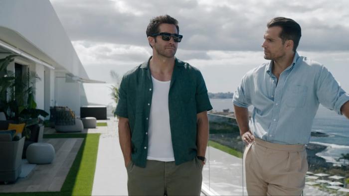 In the Grey movie: Jake Gyllenhaal and Henry Cavill walk on a sunlit patio by the sea, dressed casually, with clouds overhead