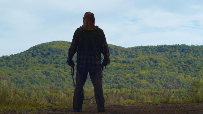 In a Violent Nature Movie: A masked killer stands, back to viewers, at the edge of a ravine, a hook and chain in his hands