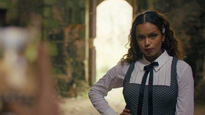 Influencers movie: Georgina Campbell in a white shirt, black ribbon tie, and overalls, right, stands in front of a doorway while outdoors