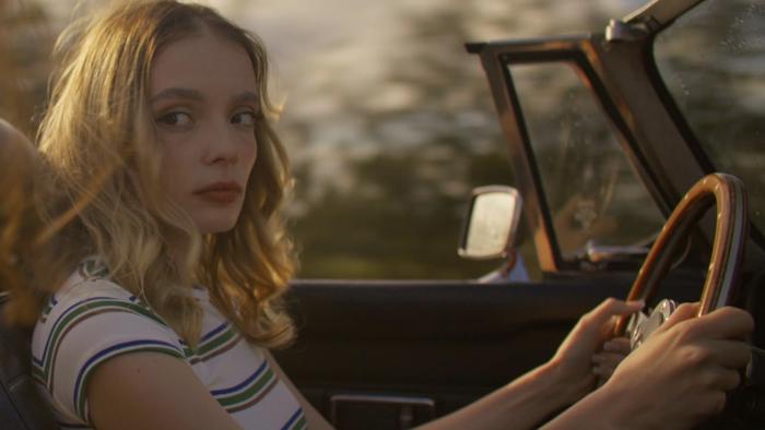 Influencers movie: White French woman with blond hair in striped grey and white shirt, left, drives a convertible with the roof down