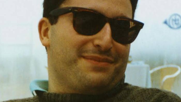 Inside the Trustor Scandal movie: Smiling man wearing sunglasses, sitting indoors.